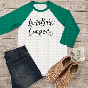 May include: A white and green baseball tee shirt with the text "LunaBabe Company" printed in black. The shirt is displayed on a wood background with blue jeans, gold glitter sneakers, and a white mug with a Christmas design.