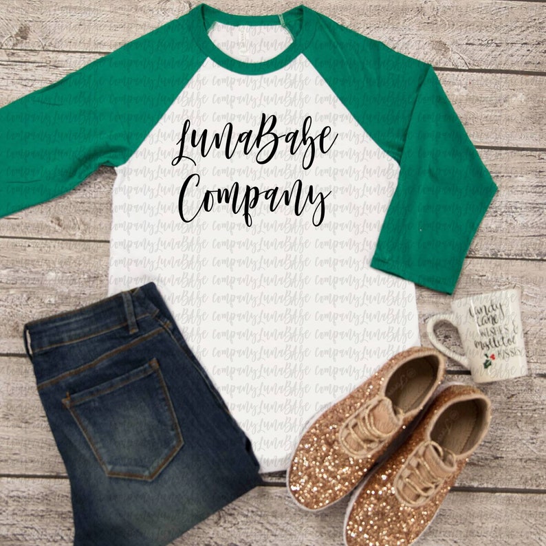 May include: A white and green baseball tee shirt with the text "LunaBabe Company" printed in black. The shirt is displayed on a wood background with blue jeans, gold glitter sneakers, and a white mug with a Christmas design.