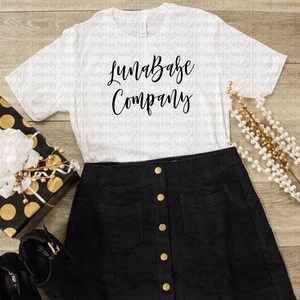May include: White t-shirt with black text that reads "LunaBabe Company". The shirt is laying on a wooden surface with a black and gold gift box, a black skirt with gold buttons, and a gold beaded garland.