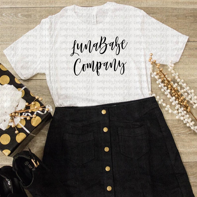 May include: White t-shirt with black text that reads "LunaBabe Company". The shirt is laying on a wooden surface with a black and gold gift box, a black skirt with gold buttons, and a gold beaded garland.
