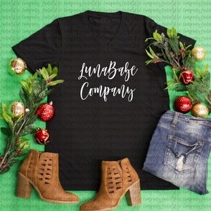 May include: A black v-neck t-shirt with the text "LunaBabe Company" printed in white. The shirt is laid flat on a green surface with red and gold ornaments and a pair of brown ankle boots.