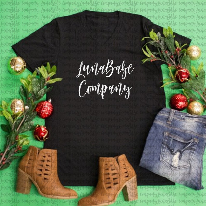 May include: A black v-neck t-shirt with the text "LunaBabe Company" printed in white. The shirt is laid flat on a green surface with red and gold ornaments and a pair of brown ankle boots.