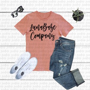 May include: A peach-coloured t-shirt with the text "LunaBabe Company" printed in black. The shirt is laid out on a white wooden surface with a pair of white trainers, blue jeans, and a small potted plant.