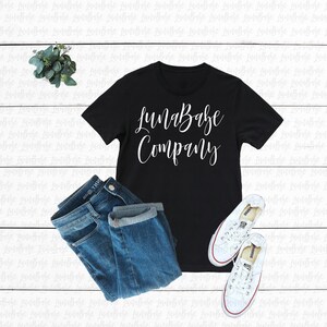 May include: Black t-shirt with white text that reads "LunaBabe Company". The shirt is laid flat on a white wooden surface with a pair of blue jeans and white sneakers.