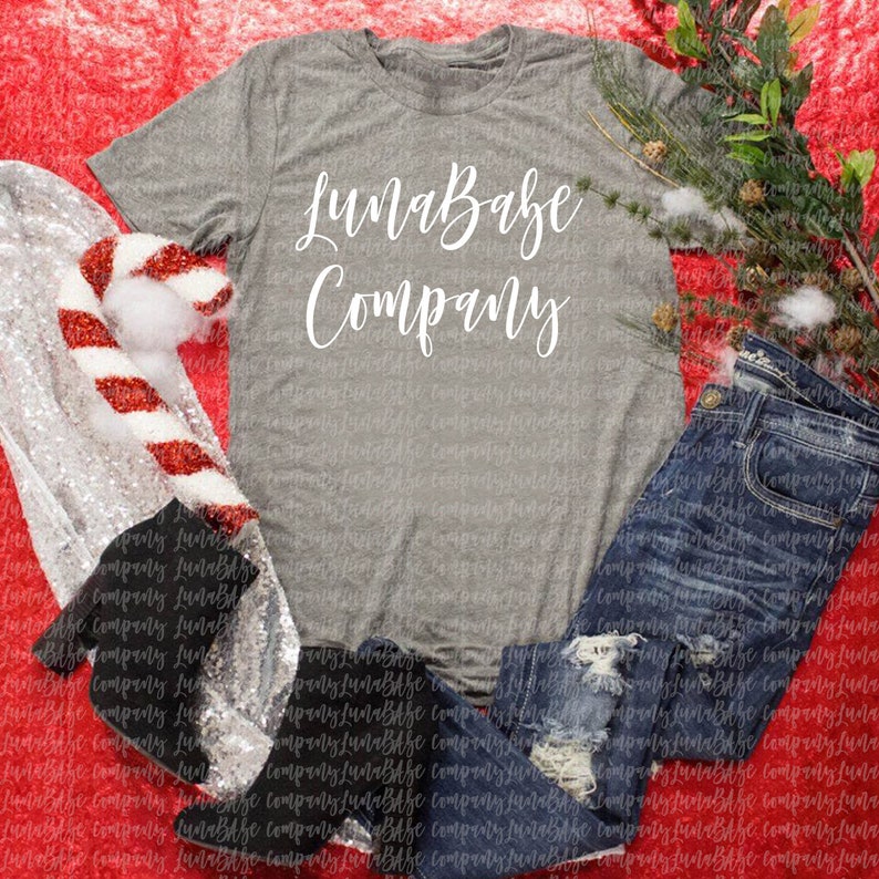 May include: A gray t-shirt with the text "LunaBabe Company" in white cursive lettering. The shirt is laid out on a red background with a silver and red striped candy cane and blue jeans.