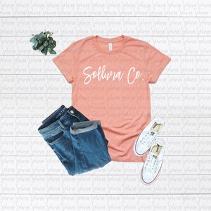 May include: A peach-coloured t-shirt with the text "Sollura Co." in white lettering. The shirt is laid flat on a white wooden surface with a pair of blue jeans and white trainers.