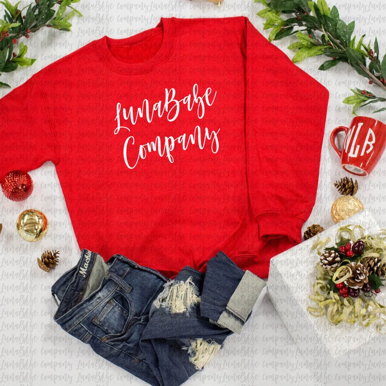 May include: A red sweatshirt with the text "LunaBabe Company" in white lettering. The sweatshirt is laid flat on a white surface with a pair of blue jeans, a red mug, and a wrapped gift box.