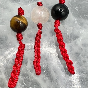 May include: Three gemstone pendants with red braided cords. The gemstones are a tiger's eye, a clear quartz, and a black onyx. The pendants are displayed against a light gray, textured background.