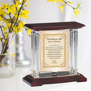May include: A mahogany wood plaque with silver columns and a glass front. The plaque features a personalized message for a new doctor, "Nick Hansen, MD Doctor of Medicine". The message includes a poem about the doctor's journey and the importance of their work.