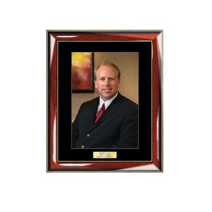 May include: A framed portrait of a man in a black suit and red tie. The frame is made of dark wood with a gold trim. The man is smiling and looking directly at the camera. The text below the portrait reads "Donner C. Lewis, Senior Vice President, Digital Wireless Network Systems."