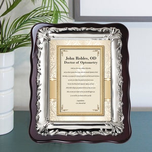 May include: A silver-framed plaque with a dark wood backing. The plaque features a gold-colored certificate with the text "John Robles, OD Doctor of Optometry" and a poem about the importance of vision.