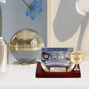 May include: A gold-colored desk set with a round, clear glass dome, a business card holder, and a gold-colored medical symbol on a stand. The business card holder has a red wood base and a gold-colored plaque with the text "Cindy Walker, CEO, Order of Dentistry, Engraved Clock by AllGiftFrames.com". The business card holder is on a white surface. The business card holder has a card with the text "Framing Achievement, Inc., www.AllGiftFrames.com, Premium Engraved Clocks, Retirement Gifts, Recognition Service Awards and Personalized Poetry Frames, info@AllGiftFrames.com".