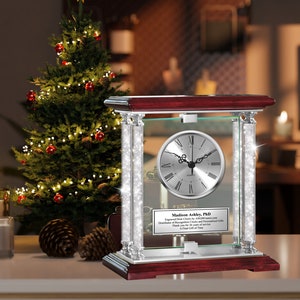 May include: A silver clock with Roman numerals on a glass base with a mahogany wood frame. The clock is surrounded by crystal-like accents. The clock has a personalized plaque with the text "Madison Ashley, PhD Engraved Desk Clock by Gifts.com Distributor of Recognition Clocks and Personalized Gifts Thank You for Your Outstanding Service A Final Gift of Time".