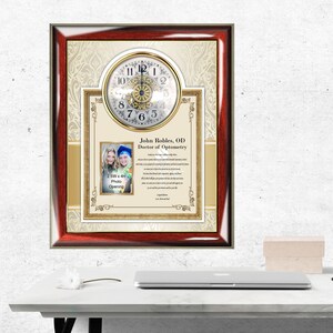 May include: A framed clock with a gold-colored clock face and a picture of two people in graduation attire. The frame is red and gold with a cream-colored background. The text "John Robles, OD Doctor of Optometry" is printed on the frame.