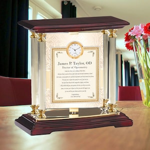May include: A mahogany and gold-tone clock with a glass front and a personalized inscription for a Doctor of Optometry. The inscription reads "James P. Taylor, OD Doctor of Optometry Inside you, there was a calling to help others. And you chose to pursue that career and attended Optometry School. And today, we prepare you for a journey in optometry and look towards the future. We want to honor your love, your compassion, your dignity and honor. You have been blessed with patience, the best care they can come. All of which will grace your partners, the firm, and home. James, we want you to know we love you and will support you. Go out and live your dreams and love your life. Love, Mom and Dad Congratulations"