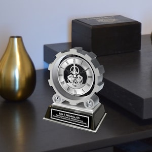 May include: A silver desk clock with a gear design and a black face. The clock has a personalized plaque that reads "Rylie L. Thibodeau, RN Engraved Desk Clocks by Awards4U.com Distributor of Recognition Clocks and Personalized Gifts".