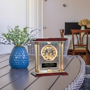 May include: A gold-toned clock with a glass front and a mahogany base. The clock has a Roman numeral face and a visible gear mechanism. A personalized plaque with the text "Madison C. McDonald" is displayed below the clock face.