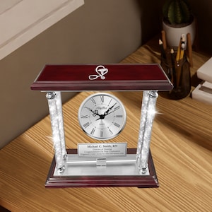 May include: A mahogany desk clock with a silver face and silver pillars. The clock has a silver stethoscope charm on top and a personalized plaque that reads "Michael C. Smith, RN Director of Nursing Congratulations On Your Graduation Engraved Desk Clocks by AllGiftFrames.com"