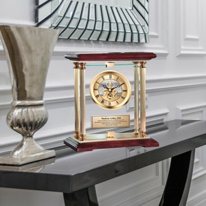 May include: A gold-toned clock with a Roman numeral face and a glass dome sits on a mahogany base with a brass plaque that reads "Madison Ashley, PhD. Engraved Desk Clock.  A symbol of your dedication, hard work and perseverance.  Thank you for the 20 years of service.  A Time Out of Time."
