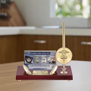 May include: A mahogany desk set with a gold pen and a gold circular base featuring a medical symbol. The base is engraved with the text "OD". The desk set is on a wooden surface. The card in the background reads "Framing Achievement, Inc. www.AllGiftFrames.com Premium Engraved Clocks, Retirement Gifts, Recognition Service Awards and Personalized Picture Frames info@AllGiftFrames.com Dr. Jane Walker, OD Doctor of Optometry Engraved Clocks by AllGiftFrames.com"