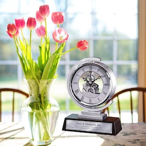 May include: A silver clock with a visible gear mechanism on a black base. The clock is engraved with the text "Cindy B. McDaniels, RN  Engraved Desk Clocks by Awards.com  Distributor of Personalized Retirement and Recognition Award Clocks". A vase with pink tulips is in the foreground.