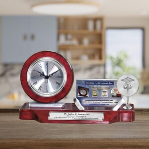 May include: A mahogany desk clock with a silver face and Roman numeral markings. The clock is on a mahogany base with a silver plaque that reads "Dr. Jadyn C. Smith, OD Doctor of Optometry ARCIPrestige Engraved Clocks and Frames". A silver plaque with the text "Framing Achievement, Inc." is on the base. A silver medical symbol is on a stand to the right of the clock.