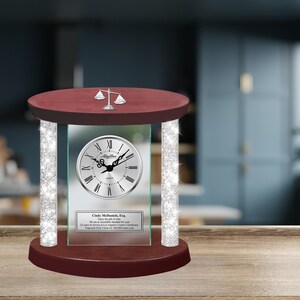 May include: A silver clock with Roman numerals on a red wooden base with a silver scale of justice on top. The clock is surrounded by a glass frame with a silver glitter border. The glass frame has a personalized inscription that reads "Cindy McMannis, Esq. We are so proud to celebrate the dedication of your 25 years of service to the legal profession. Engraved Desk Clocks for All Occasions www.GiftIdeas.com".