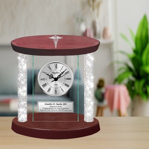 May include: A red wood base with a glass clock face and a silver medical symbol on top. The clock face has Roman numerals and a silver frame. The glass is surrounded by a silver glitter border. The text on the plaque reads "Jennifer D. Smith, RN Registered Nurse Congratulations on Your Graduation Engraved Desk Clock by AllGiftFrames.com".