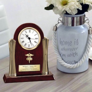 May include: A mahogany desk clock with gold accents and a white face. The clock has a medical symbol and the text "Michael C. Walker, OD Doctor of Optometry Congratulations On Your Graduation Engraved Desk Clocks by AllGiftFrames.com".