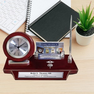 May include: A mahogany desk clock set with a silver clock face, a silver pen holder, and a silver business card holder. The clock face has Roman numerals and the set has a personalized plaque that reads "Ryder L. Thomas, OD Doctor of Optometry Engraved Desk Clocks by AllGiftFrames.com".