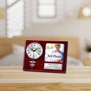 May include: A mahogany wood desk clock with a silver clock face and a silver medical symbol. The clock has a 3x3 inch photo frame with a woman in it. The text on the plaque reads "Cindy E. Thomas, OD. Enjoy the gift of health... We are so incredibly thankful for all of your years of service at New York Eye Care.  Forever grateful, Cindy by AllGiftFrames.com".