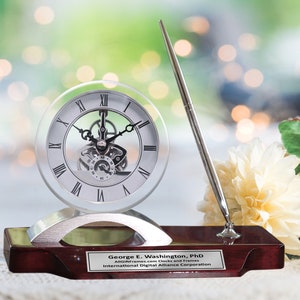 May include: A silver desk clock with a clear glass face and a silver pen on a mahogany base. The clock has a visible gear mechanism and Roman numeral markings. The base has a silver plaque with the text "George E. Washington, PhD AllGiftFrames.com Clocks and Frames International Digital Alliance Corporation".