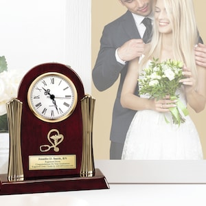 May include: A mahogany wood clock with gold accents and a stethoscope and heart symbol engraved on the front. The clock has a white face with black Roman numeral hour markers and black hands. The clock is on a white surface with a framed photo of a bride and groom in the background. The plaque on the clock reads "Jennifer D. Smith, RN Registered Nurse Congratulations On Your Graduation Engraved Desk Clock by AddallGifts.com".