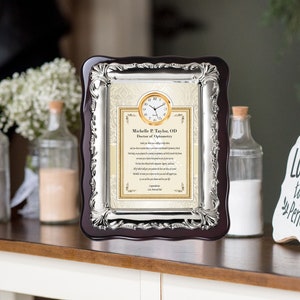 May include: A framed certificate with a clock at the top, featuring the name "Michelle P. Taylor, OD Doctor of Optometry." The frame has a dark wood border with silver accents. The background is a cream color with decorative elements.