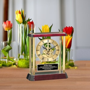 May include: Gold-toned clock with a visible gear mechanism, enclosed in a glass case with a mahogany base. The clock has Roman numeral markings and a personalized plaque that reads "Madison James Engraved Desk Clocks by AllGiftIdeas.com Distributor of Personalized Gifts Thank you for 20 years of service A Gift of Time".