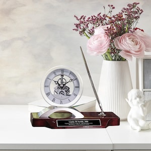 May include: A silver desk clock with a visible gear mechanism and a matching pen stand on a mahogany base. The base has a personalized plaque with the text "Linda M Smith, MD Engraved Desk Clock & Pen Set by AMZGEAR Distributor of Recognition Clocks".