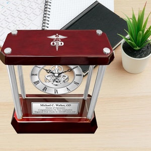 May include: A mahogany wood desk clock with a silver face and a silver open gear mechanism. The clock has a silver plaque with the text "Michael C. Walker, OD Doctor of Optometry Congratulations On Your Graduation Engraved Desk Clocks by AltFrames.com".