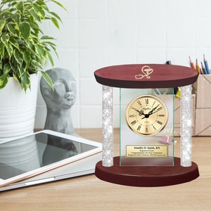 May include: A mahogany wood desk clock with a glass front and a gold-colored clock face. The clock is surrounded by a silver glitter border. The glass front has a personalized inscription that reads "Jennifer D. Smith, RN Registered Nurse Congratulations On Your Graduation Engraved Desk Clocks By AwardsandThings.com".