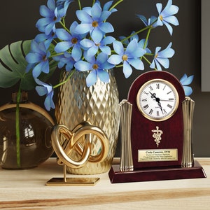 May include: A mahogany clock with gold accents and a white face. The clock has a plaque with the text "Cindy Cameron, DVM, Doctor of Veterinary Medicine, Congratulations on Your Retirement,  Engraved Desk Clocks by AddGifts.com". The clock is on a wooden surface with a gold knot sculpture, a glass vase with blue flowers, and a clear glass vase with a green stem.