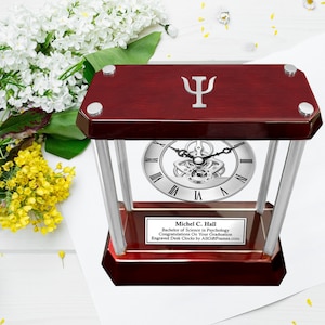 May include: A mahogany wood desk clock with a silver face and a silver gear mechanism. The clock is engraved with the text "Michel C. Hall, Bachelor of Science in Psychology, Congratulations On Your Graduation, Engraved Desk Clocks by AllGiftFrames.com". The top of the clock has a silver Greek letter Psi symbol.