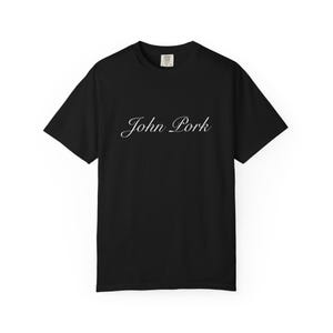 May include: Black t-shirt with the white cursive text "John Pork" across the chest. The shirt has short sleeves and a crew neck. The shirt is made of a soft material.