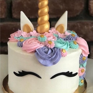 May include: A white unicorn cake decorated with pink, purple, and blue frosting flowers. The cake has a gold horn and white ears. The cake is on a gold cake board.