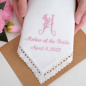 May include: White handkerchief with lace trim, embroidered with a pink floral initial "H", the text "Mother of the Bride" and the date "April 8, 2022".