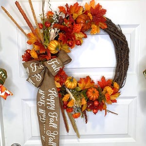May include: A fall-themed wreath with a burlap ribbon that says "Happy Fall Y'all". The wreath is made of grapevine and is decorated with faux leaves, pumpkins, and berries in shades of orange, red, and yellow.