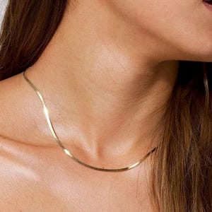 May include: A gold-toned herringbone chain necklace resting against the collarbone. The necklace features a flat, flexible, and polished metal link design that sits close to the neck, creating a smooth and reflective surface across the entire chain.