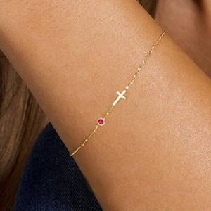 May include: A gold chain bracelet with a small cross charm and a red gemstone accent.