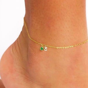 May include: A delicate gold anklet featuring a small green gemstone and the letter "E" charm. The anklet is worn on an ankle, highlighting the jewellery's design. The chain is thin and elegant, suitable for everyday wear.