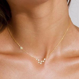 Puede incluir: Un delicado collar de oro con un diseño de constelación y pequeños acentos brillantes. El collar incluye una sola letra inicial. La joya se lleva en el cuello de una persona, contra una parte superior blanca.