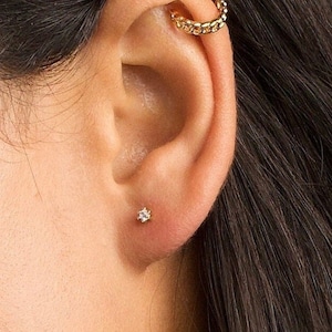 May include: A close-up of a person's ear with two gold earrings. The earring in the upper ear is a small hoop with a chain design. The earring in the lower ear is a small stud with a single clear stone.