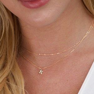 May include: A delicate gold necklace with three strands. The shortest strand features a small pendant with the letter "K". The necklace is worn against a white top. The model has blonde hair.